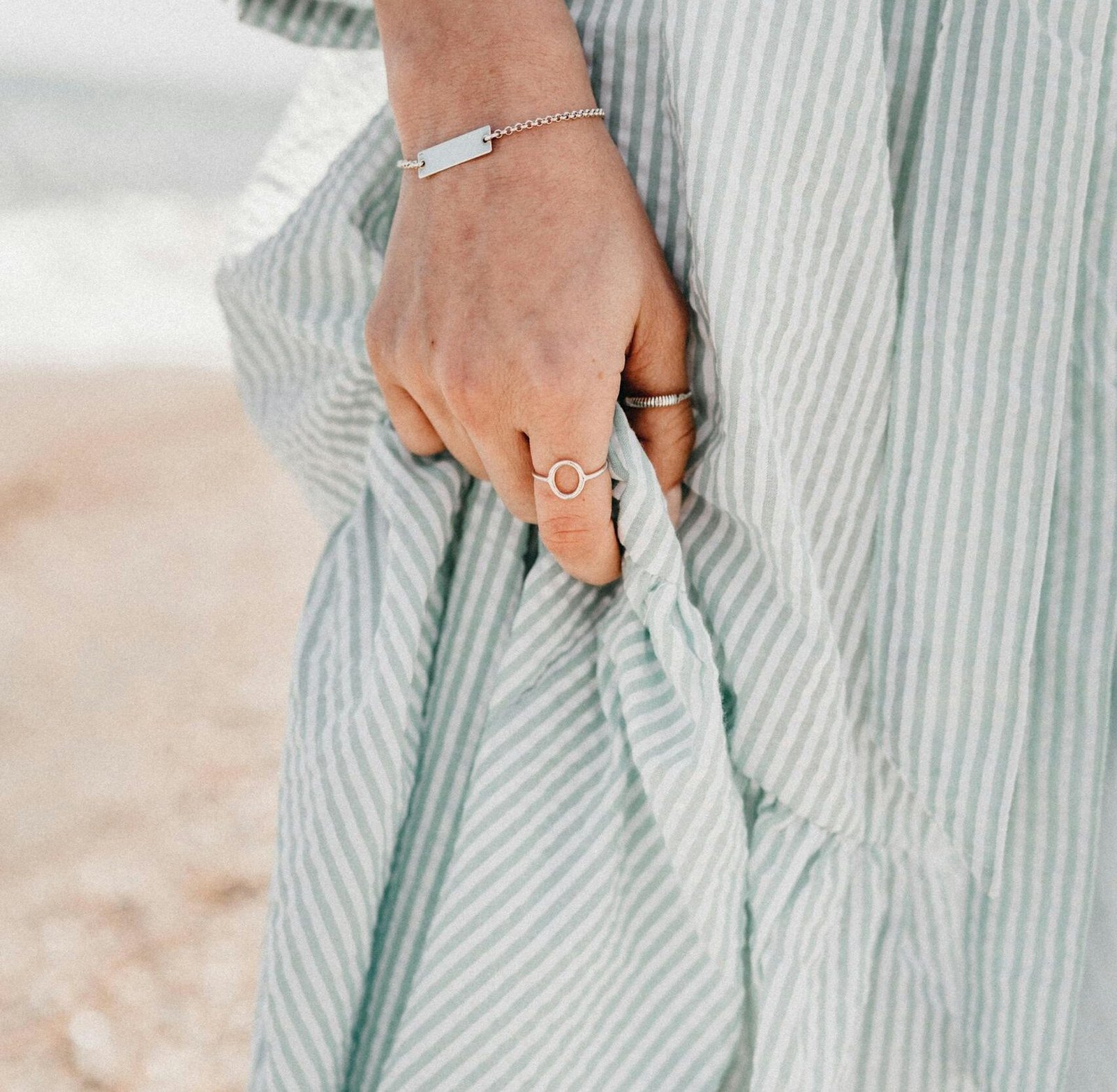 Female hand with ring and bracelet holding cotton light dress on background of sandy seashore in sunny day