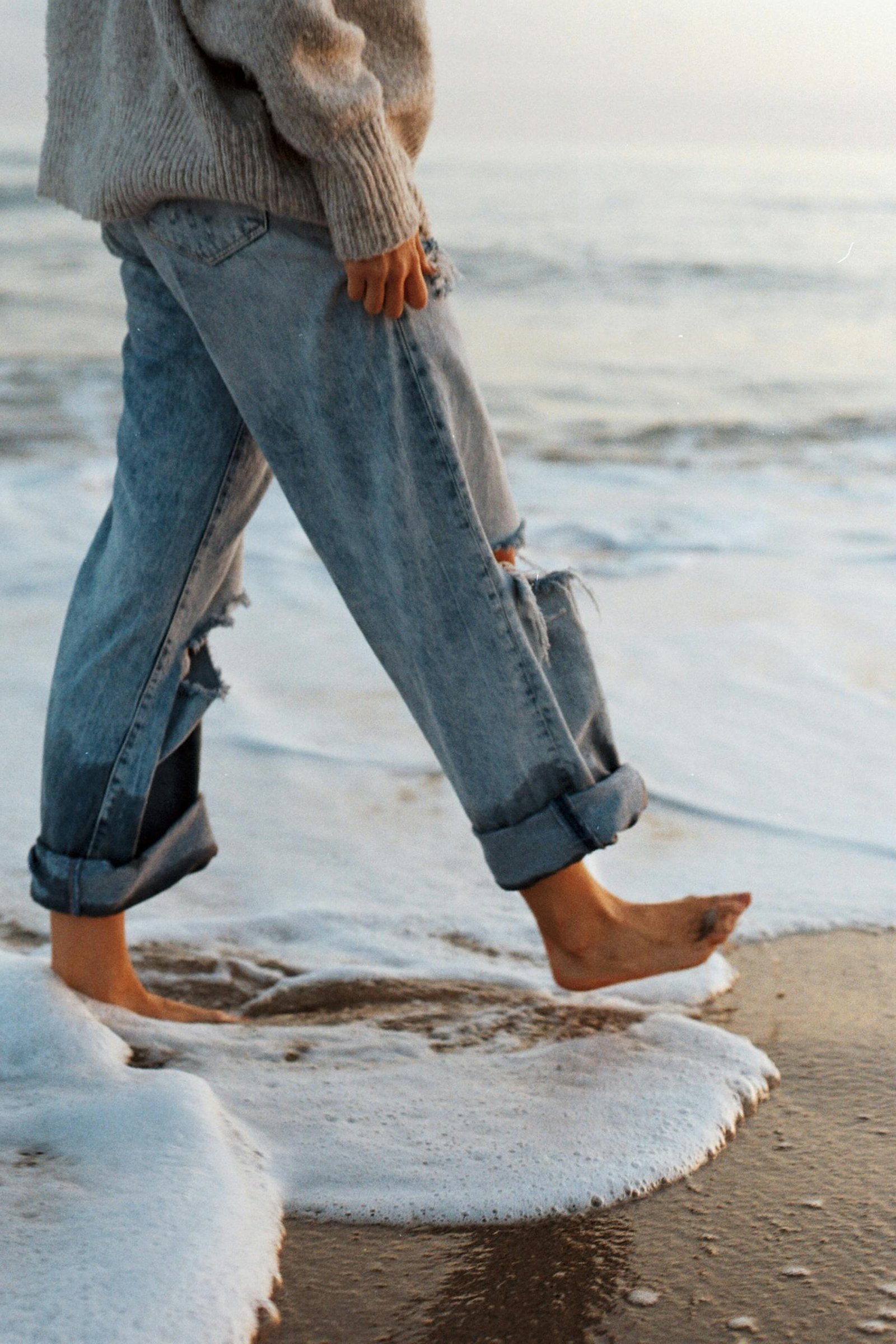 Side view of crop unrecognizable barefooted female tourist in stylish jeans and sweater walking on sandy beach washing by foamy waves of ocean during holidays